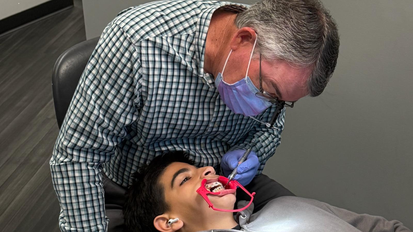 Orthodontist examining a teenage boy’s braces during a dental appointment, using a dental tool in a clinic setting.