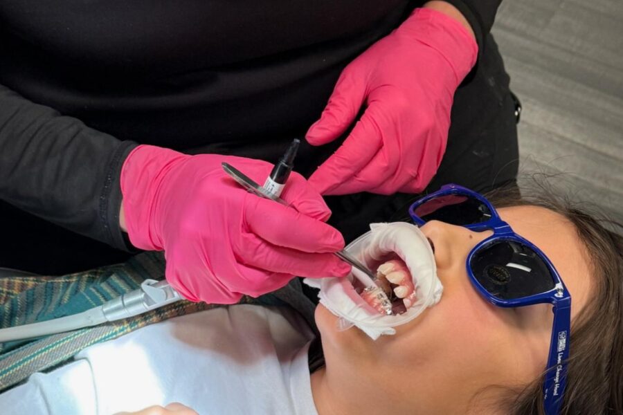 Dental assistant wearing pink gloves adjusts a young girl’s braces while she wears protective sunglasses during an orthodontic procedure.